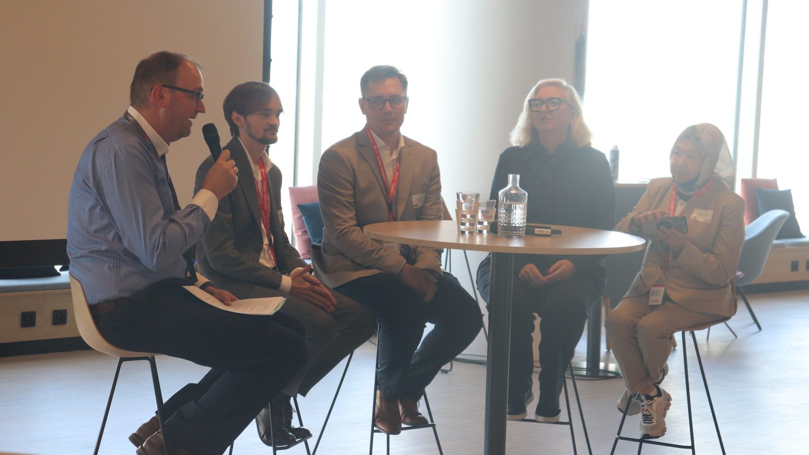Five panellists on stools around a high table, one speaking into a microphone, in the conference room at Brussels Airport