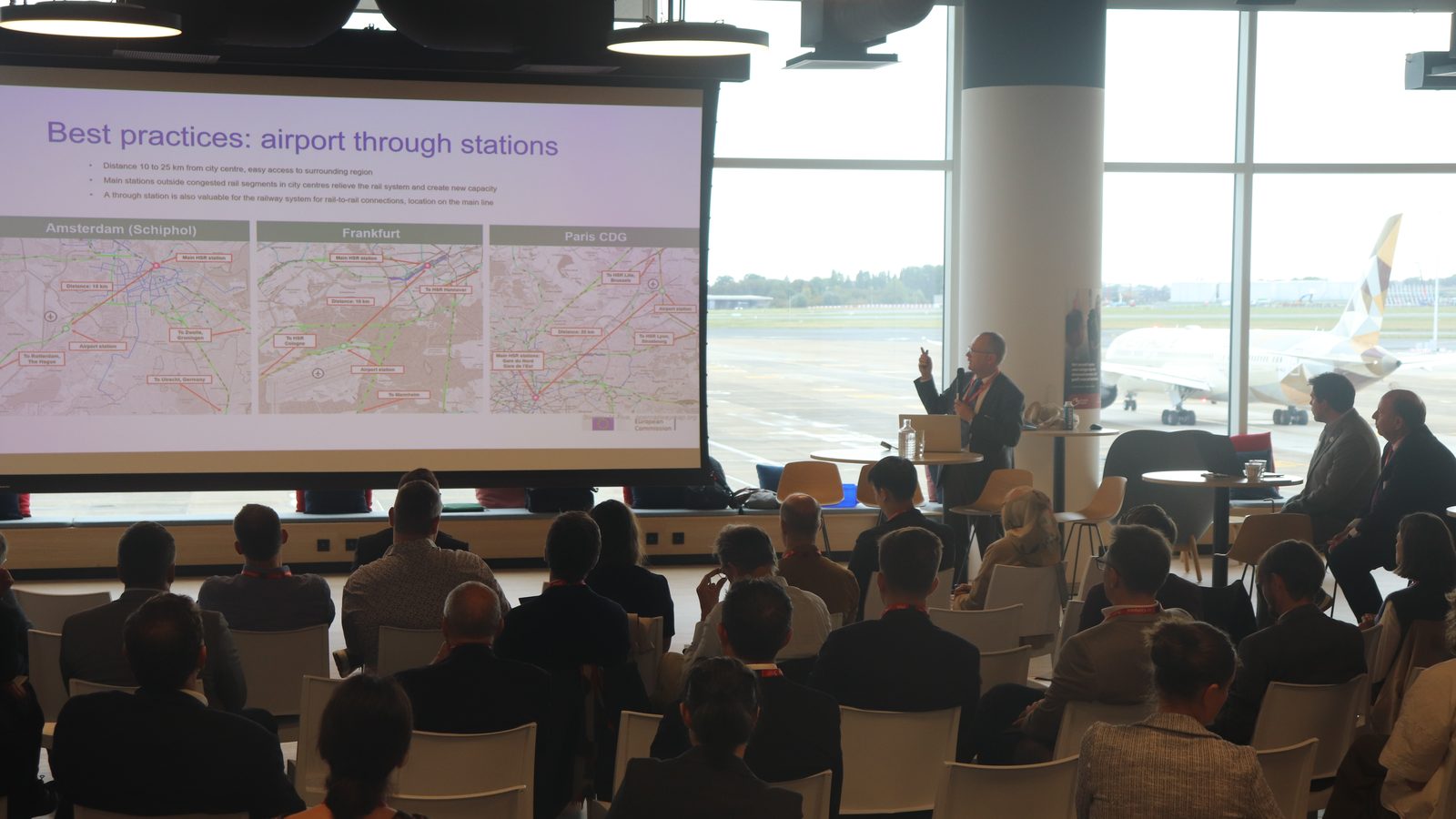 Delegates listen to a presentation on airport through stations, with aircraft on the apron visible through the windows of the conference room