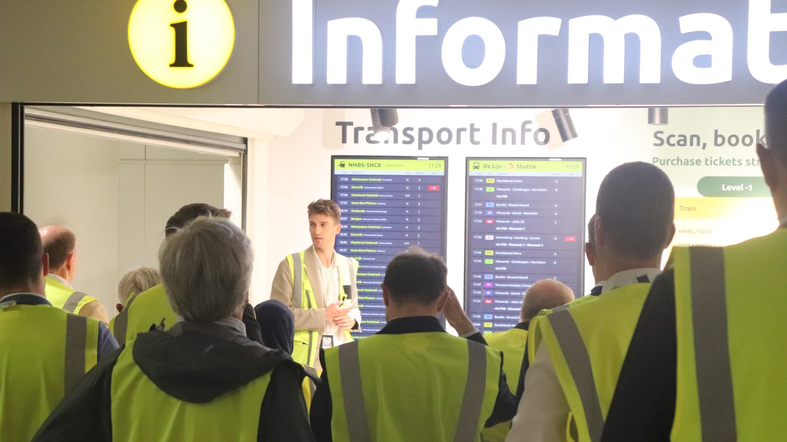 Delegates in high-visibility vests gathered at the Transport Info desk in Brussels Airport, under train departure screens