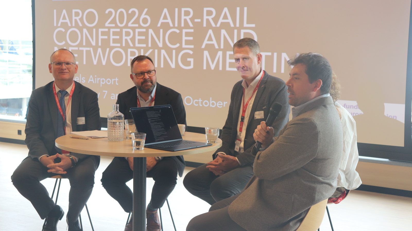 Four panellists on stools at a high table in front of the IARO 2026 Air-Rail Conference title screen at Brussels Airport