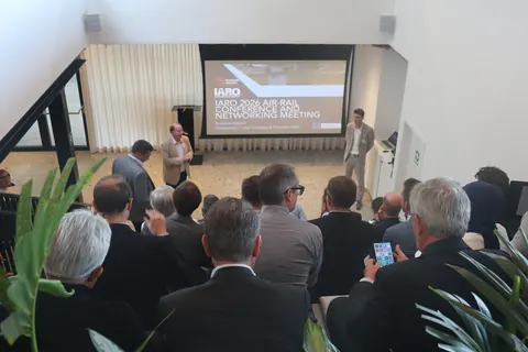 Delegates seated facing a screen showing the title slide, IARO 2026 Air-Rail Conference and Networking Meeting, with two people standing at the front