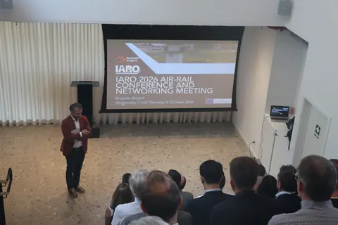A man in a red jacket speaks to the audience in front of the conference title slide