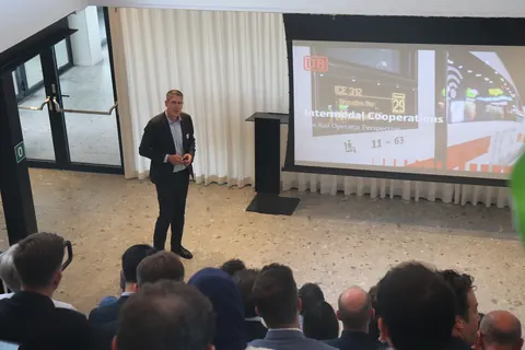 Sebastian Rieckesmann presenting beside a Deutsche Bahn slide on intermodal cooperations, with an ICE train departure display