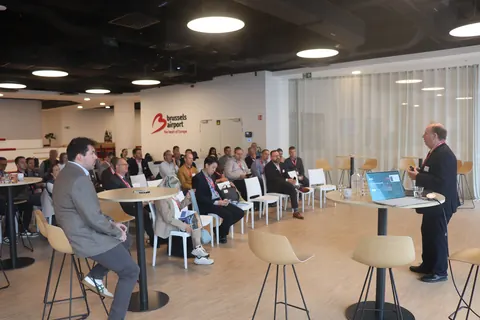 A speaker standing at a high table with a laptop addresses delegates seated in rows, under the Brussels Airport logo
