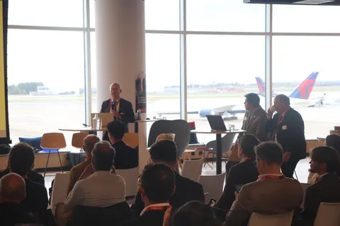 A speaker at a lectern in front of floor-to-ceiling windows, with aircraft on the apron outside