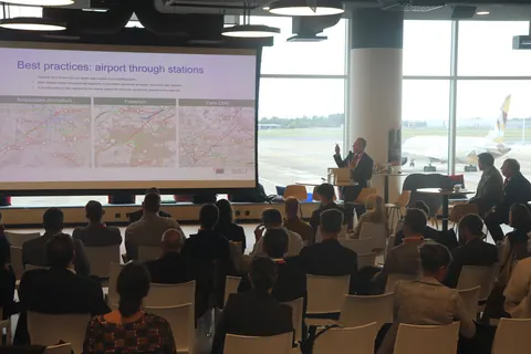 Delegates listen to a presentation on airport through stations, with aircraft on the apron visible through the windows