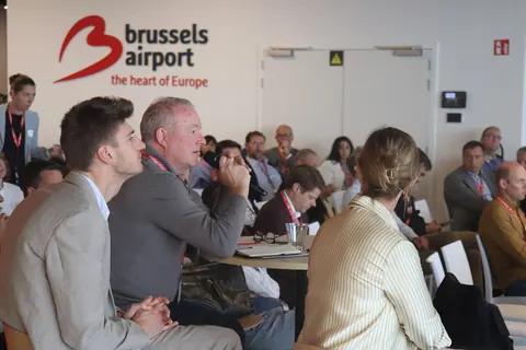 A delegate asks a question into a microphone, seated among the audience under the Brussels Airport logo