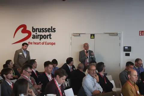 A delegate stands with a microphone to ask a question, among seated delegates under the Brussels Airport logo