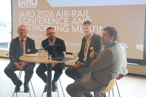 Four panellists seated at a high table in front of the conference title slide, one speaking into a microphone