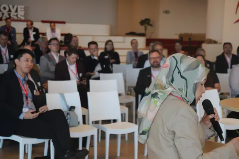 A delegate speaks into a microphone in the foreground, with other delegates seated behind