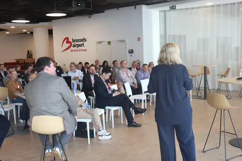 A speaker in a navy suit addresses delegates seated around the conference room