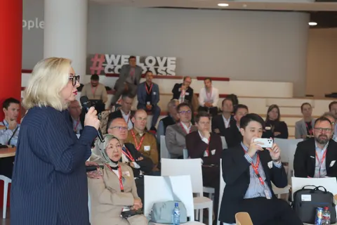 A speaker with a microphone stands beside seated delegates, one of them taking a photograph on a phone