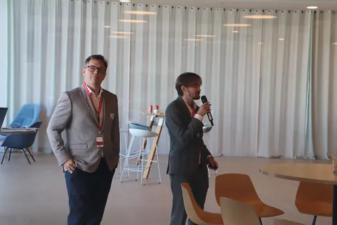 Two speakers in front of white curtains, one speaking into a microphone