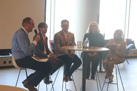 Five panellists on stools around a high table, one speaking into a microphone, in the conference room at Brussels Airport