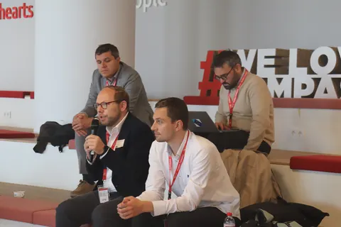 Delegates seated on a bench in front of a 'We love' sign, one speaking into a microphone