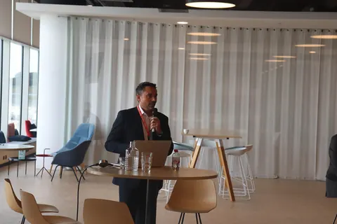 A speaker with a microphone stands behind a high table with a laptop