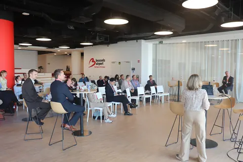 A speaker standing in the foreground addresses delegates seated in the conference room