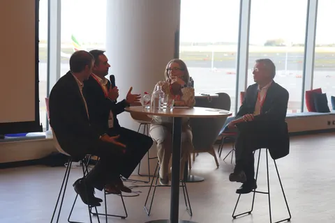 Four panellists seated around a high table in front of windows onto the airfield, one gesturing while speaking