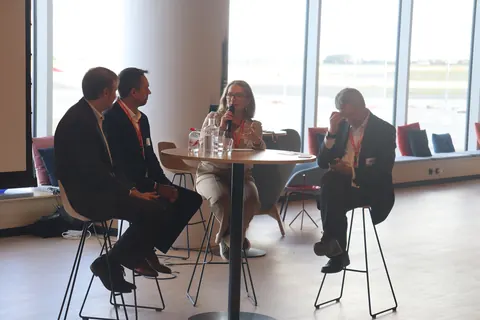 Four panellists seated around a high table in front of windows onto the airfield, one speaking into a microphone